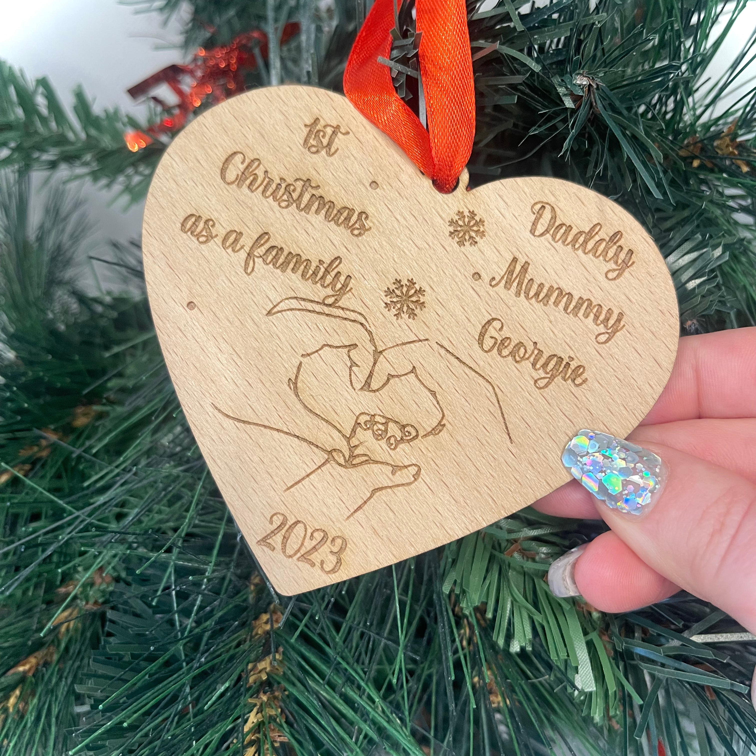Custom Heart Shaped Christmas Bauble: Crafted from 4mm beech veneer wood, this personalized ornament commemorates '1st Christmas as a Family' with engraved names. Adorned with intertwined hands, year, and a red ribbon, it adds a heartwarming touch to any tree.