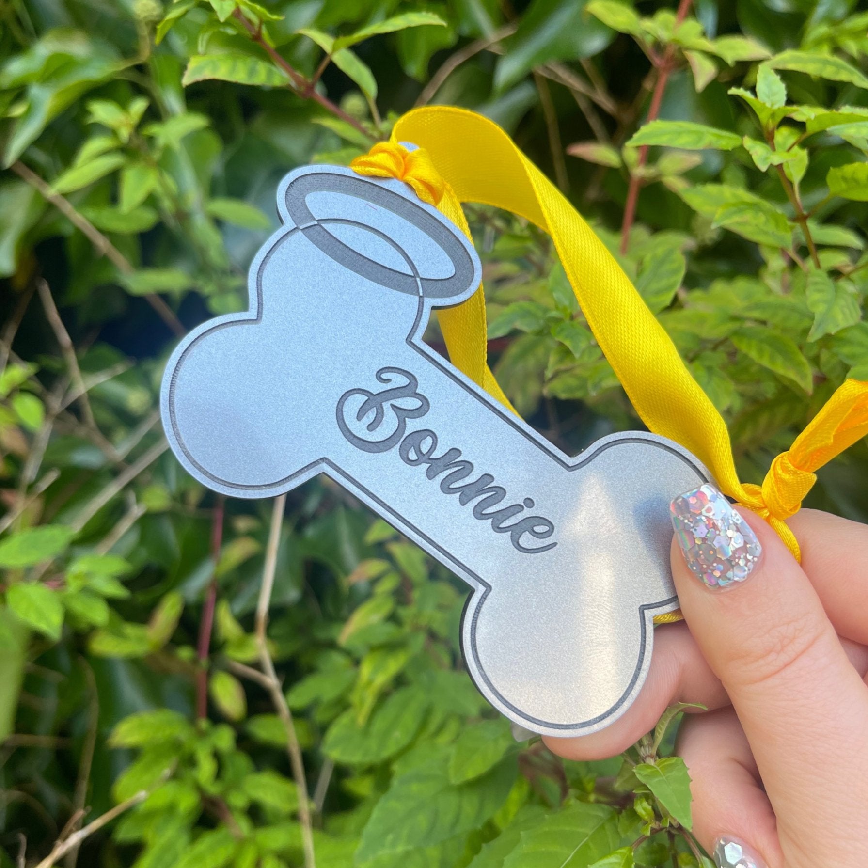 Dog Remembrance Hanging Bone Keepsake: An acrylic bone-shaped memento suspended by a yellow ribbon, featuring black engraving and a halo design. Preserve the memory of your dear dog for years to come with their personalized name on this 3mm thick keepsake.
