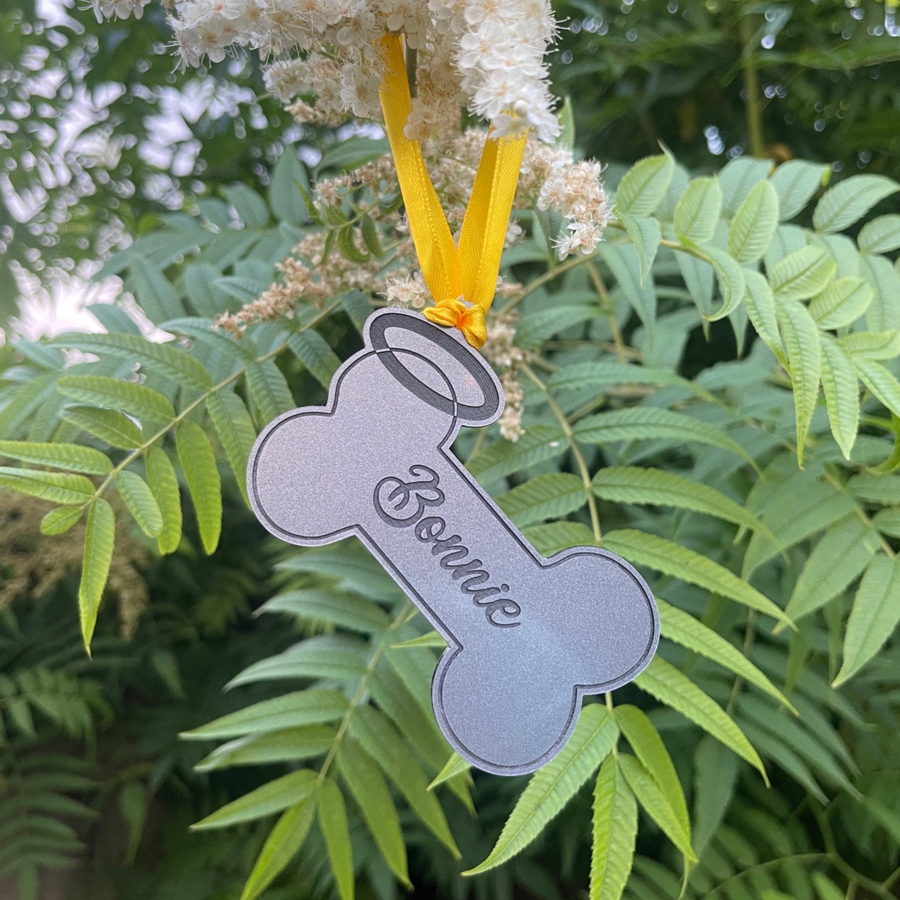 Personalized Dog Remembrance Hanging Bone with Halo: A yellow ribbon hangs from a heart-shaped hole at the top of a 3mm thick acrylic bone. The bone is engraved with a black name and surrounded by a halo. This memorial keepsake is a heartfelt way to remember a cherished dog, personalized and enduring.