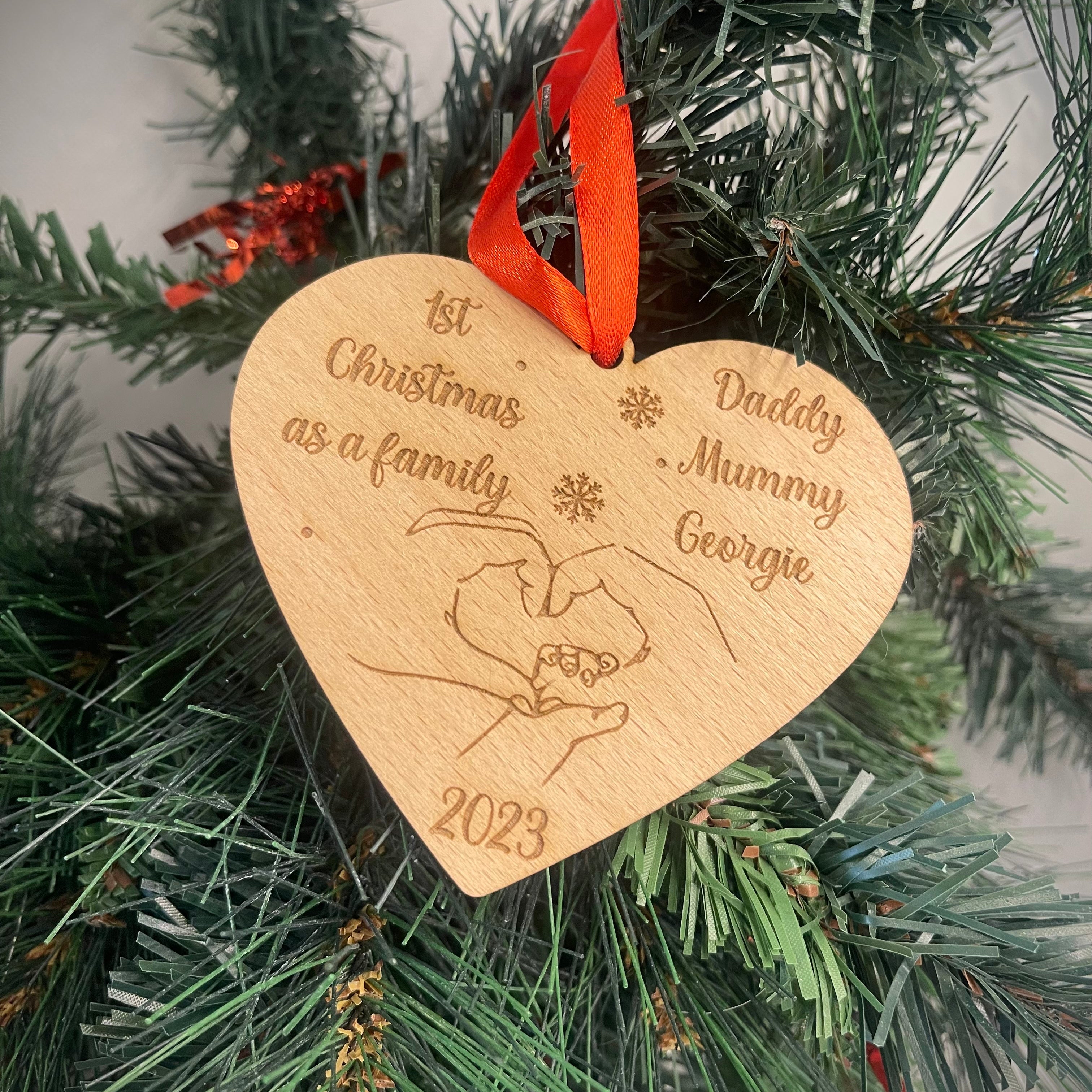 Personalized Wooden Heart Christmas Bauble: Cherish memories with our heart-shaped beech veneer ornament. Engraved '1st Christmas as a Family' with names, featuring intertwined hands, year, and a red ribbon. A heartfelt addition to your festive decor.