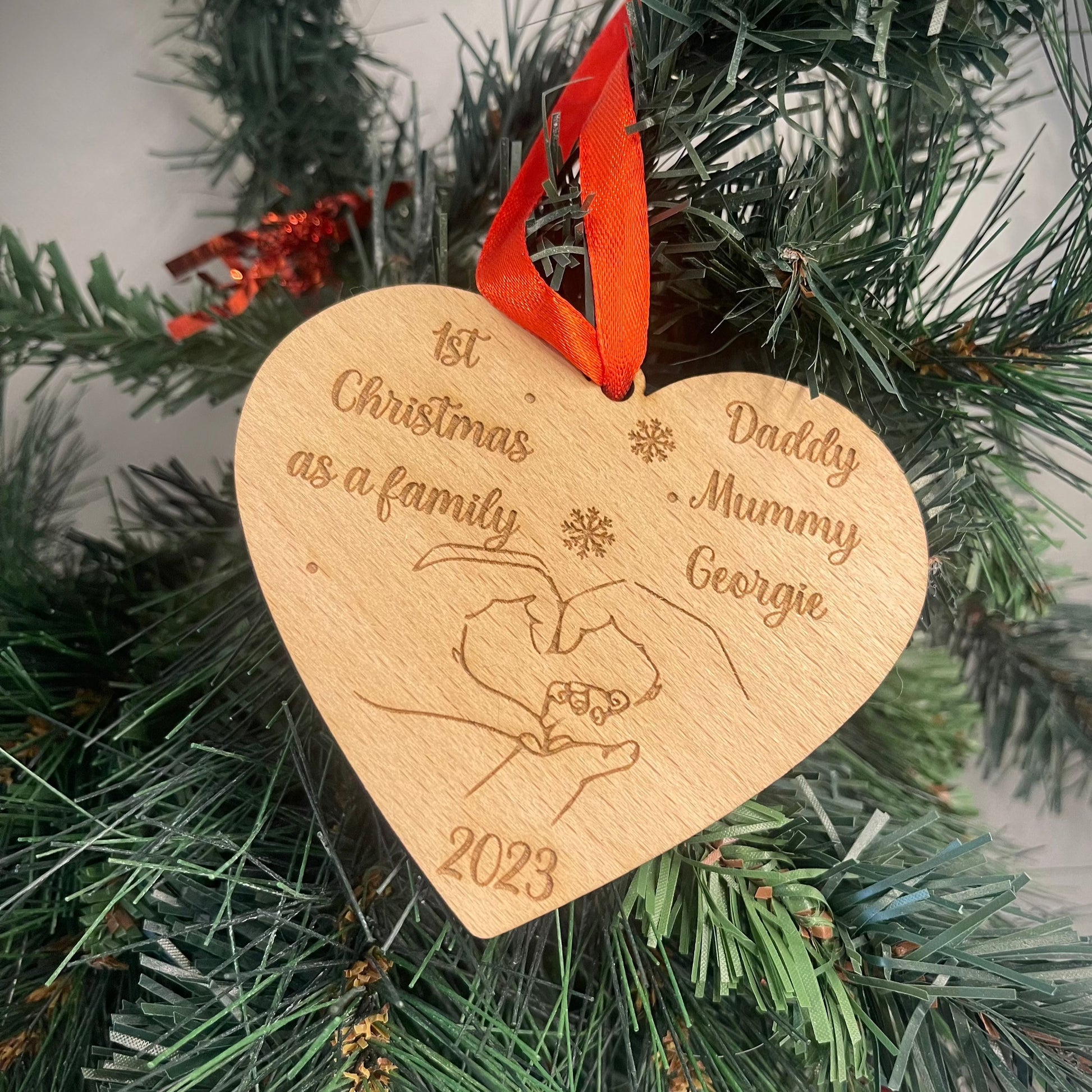 Personalized Wooden Heart Christmas Bauble: Heart-shaped beech veneer wood ornament engraved with '1st Christmas as a Family' and personalized names. Features intertwined hands, year, and red ribbon. Festive and sentimental addition to your holiday tree.