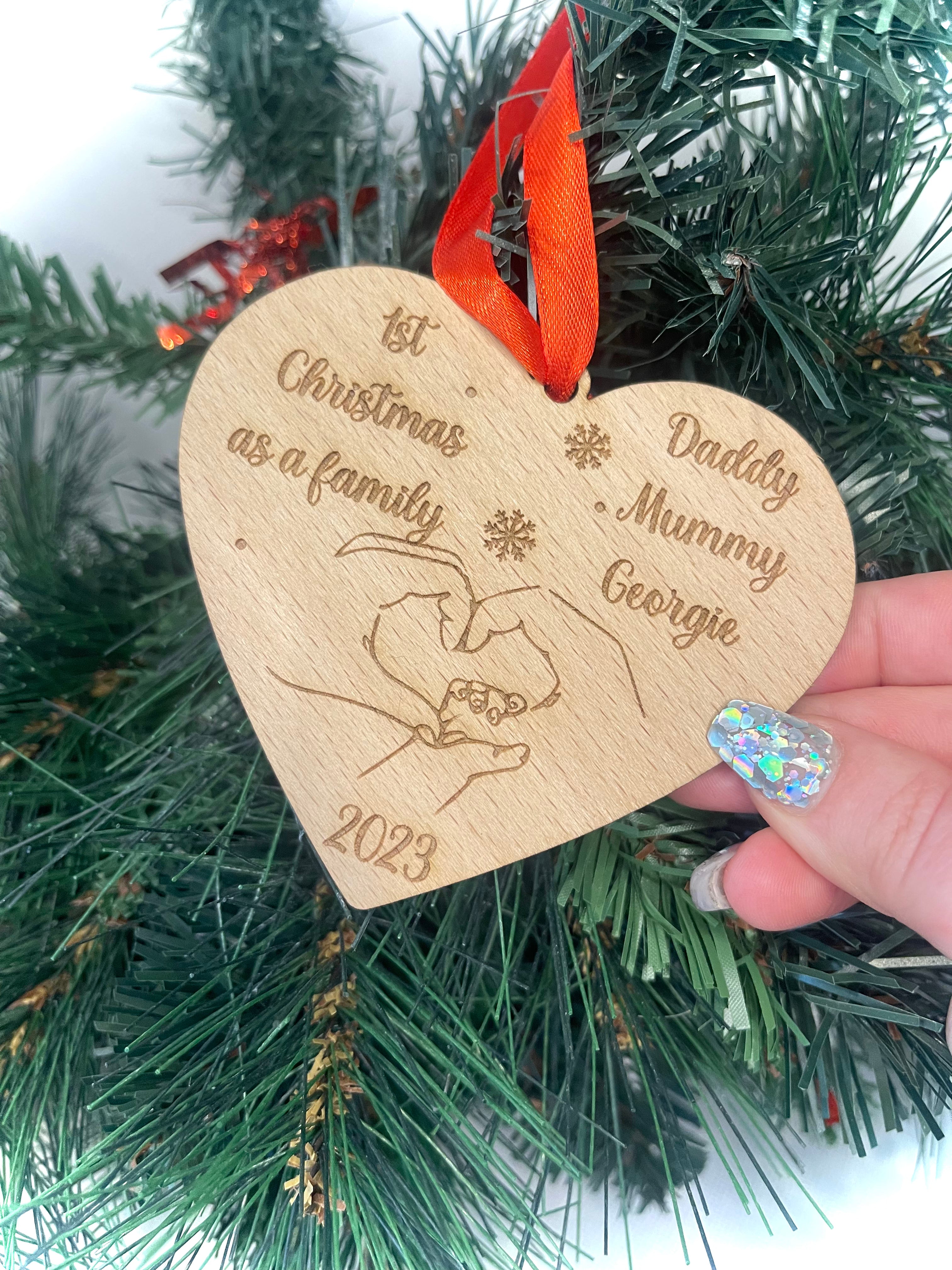 Custom Heart Shaped Christmas Bauble: Crafted from 4mm beech veneer wood, this personalized ornament commemorates '1st Christmas as a Family' with engraved names. Adorned with intertwined hands, year, and a red ribbon, it adds a heartwarming touch to any tree.