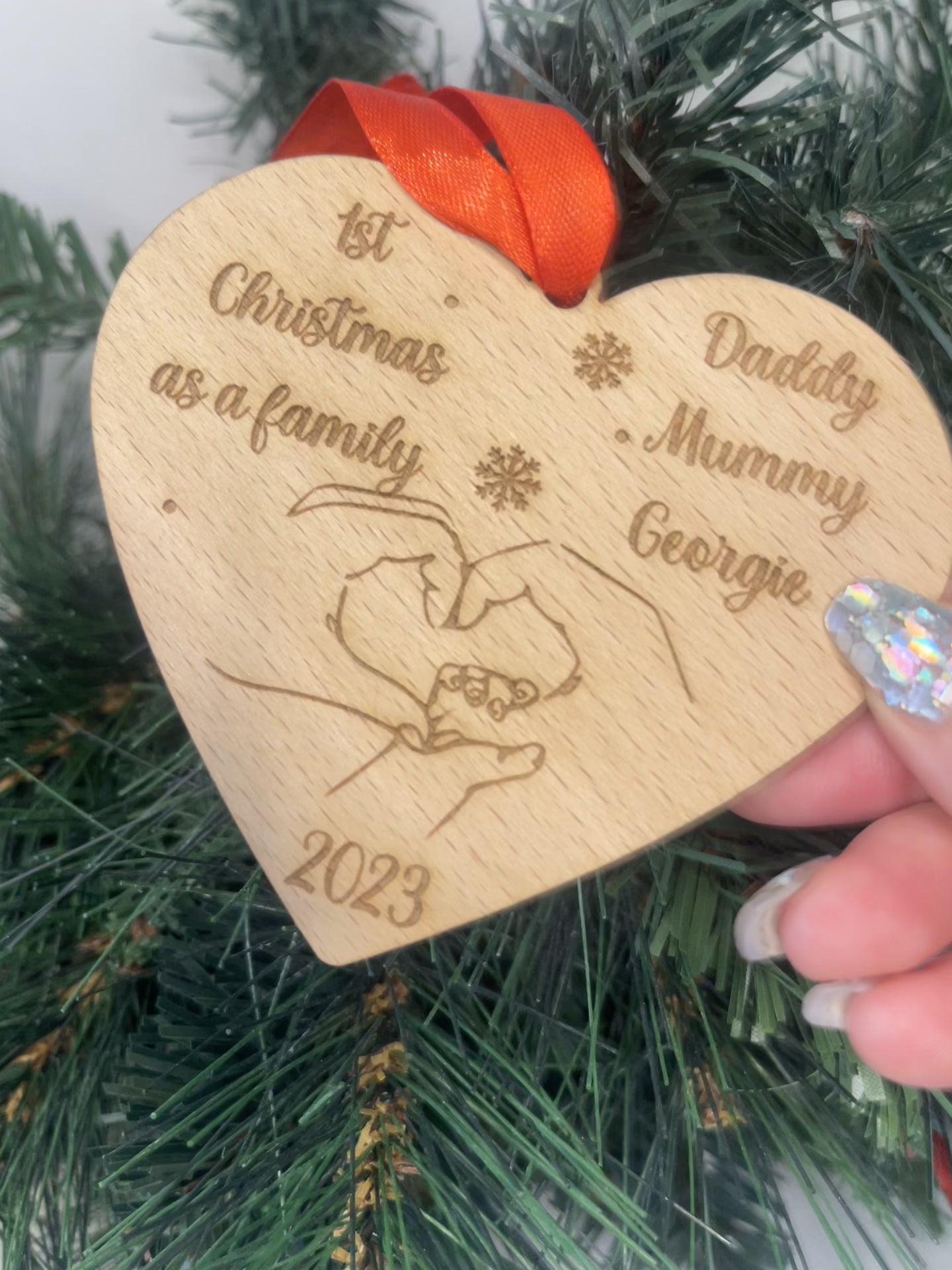 "Video: Explore Our Personalised Wooden Heart Shaped Christmas Bauble. Crafted from 4mm beech veneer wood, this ornament is engraved with '1st Christmas as a Family' and your chosen names. Watch as the heartwarming image of intertwined hands, year, and red ribbon come to life, adding a festive touch to any tree.