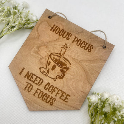 Product Image: Embrace the Halloween spirit with our captivating hanging sign. The design boasts a charmingly eerie coffee cup and ghost spoon illustration, accompanied by the phrase 'Hocus pocus, I need coffee to focus' for a festive touch. Hang it indoors to infuse your space with the enchantment of the spooky season.