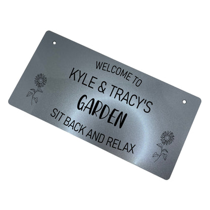 Alt text: Personalised Garden Sign - Silver Acrylic Description: A personalized garden sign in silver finish, made from high-quality 3mm thick acrylic. Engraved with "Welcome to [Your Name's] Garden" at the top, "Garden" in the middle, and "Sit Back and Relax" at the bottom. Adorned with two laser-engraved sunflowers. Weather-resistant and equipped with two 5mm holes and black twine for hanging.