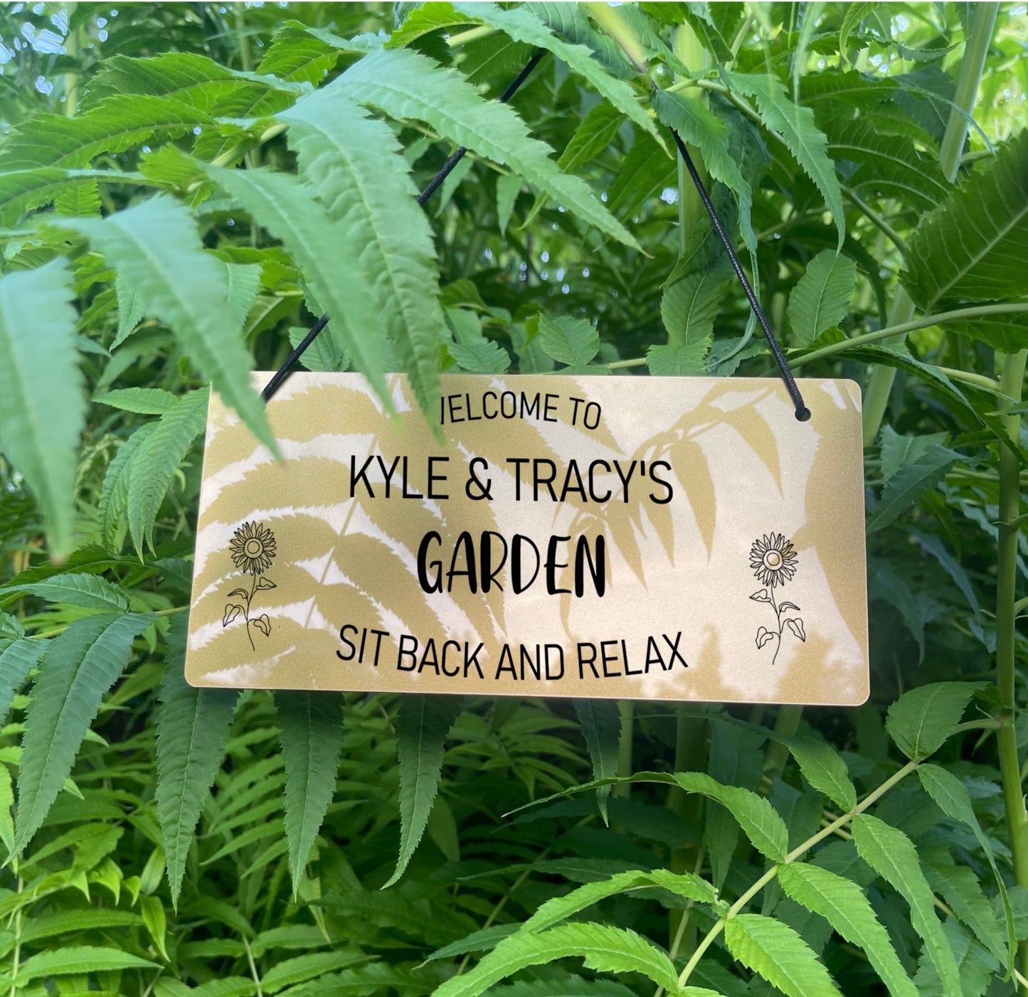 Alt text: Personalised Laser Engraved Garden Sign - Gold Finish Description: An elegant gold garden sign expertly crafted from durable 3mm thick acrylic. Features engraved text "Welcome to [Your Name's] Garden" at the top, "Garden" in the middle, and "Sit Back and Relax" at the bottom. Decorated with two laser-engraved sunflowers on either side. Comes with two 5mm holes and black twine for easy hanging.