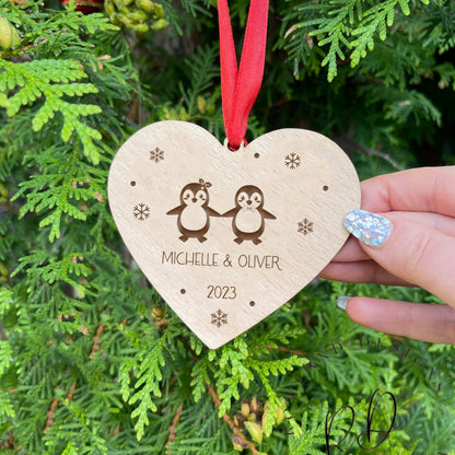 Custom Wooden Heart-Shaped Christmas Bauble with Interlocking Penguins, Snowflakes, Personalized Engravings, and Red Ribbon. Perfect Holiday Decor and Gift for Couples. 🎄🎅