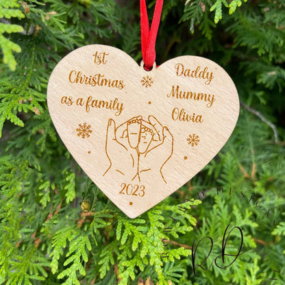 Personalized Wooden Heart-Shaped Bauble hanging on a Christmas tree branch. Engraved with '1st Christmas as a Family' and intertwined hands design, made from premium 4mm beech veneer wood. Adorned with a festive red ribbon